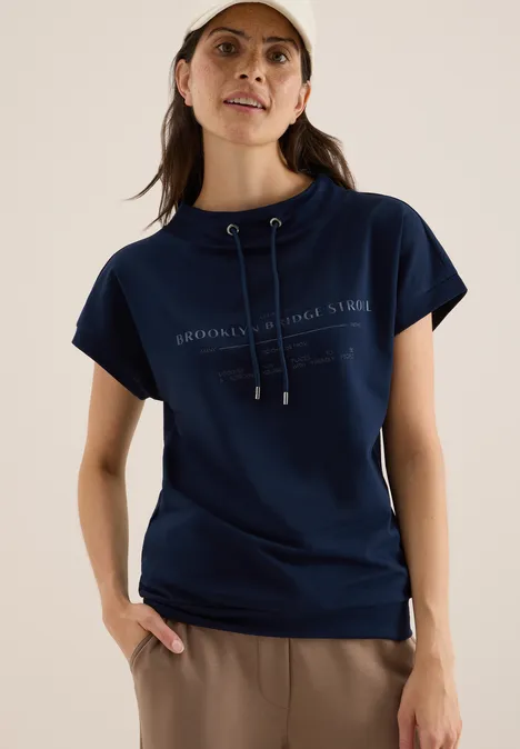Frau mit dunkelblauem Shirt und heller Kappe steht lächelnd vor neutralem Hintergrund.